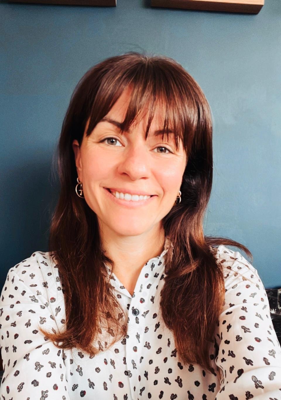 A woman with shoulder-length brown hair is smiling and taking a selfie. She is wearing a white shirt with small butterfly and flower patterns and earrings. The background is a solid dark blue wall.