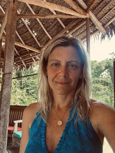 Woman with blond hair wearing a blue sleeveless top taking a selfie under a thatched roof structure in a lush, green outdoor setting.