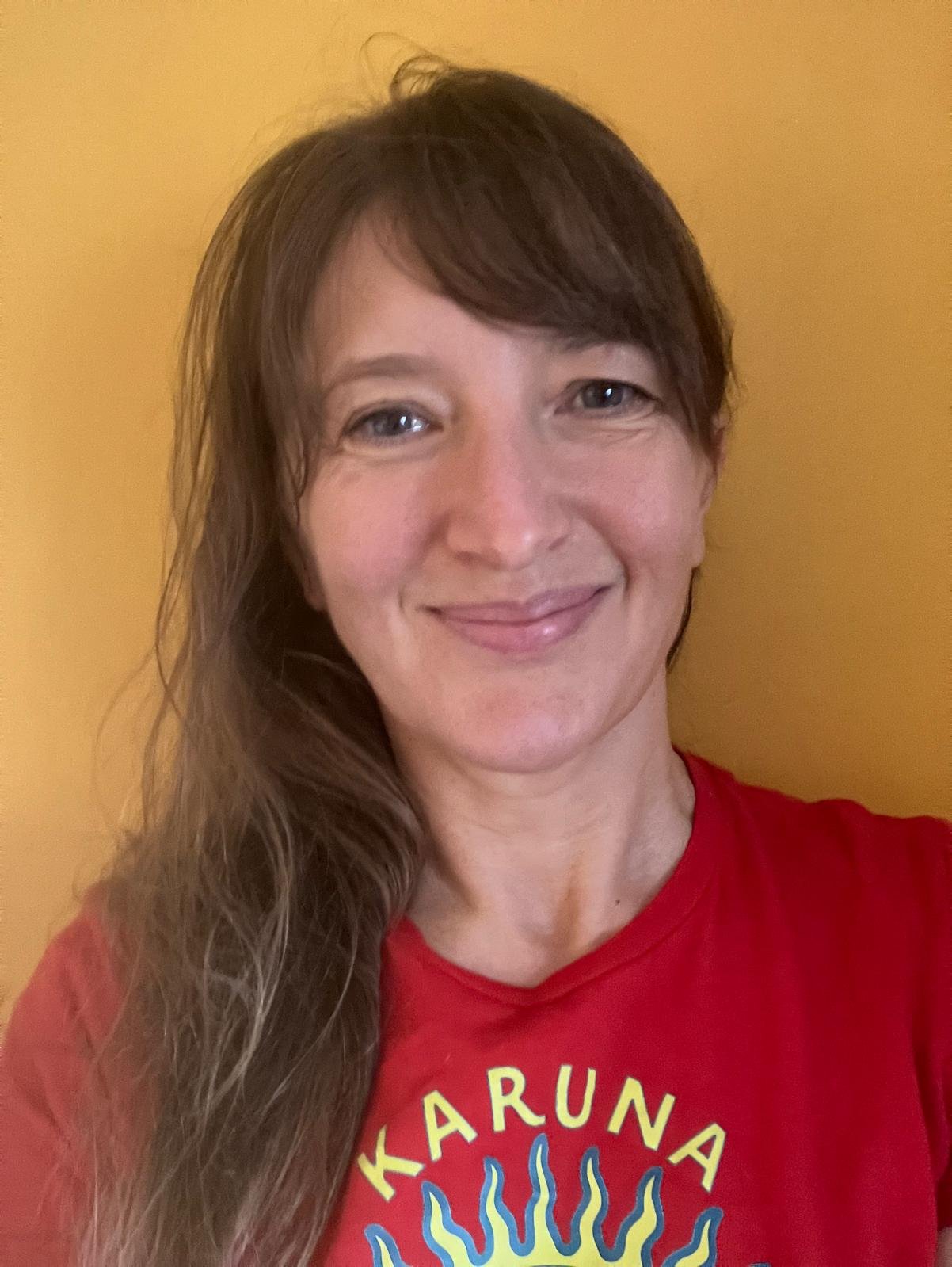 A woman with long brown hair smiling in front of a yellow background, wearing a red t-shirt with yellow and blue design.