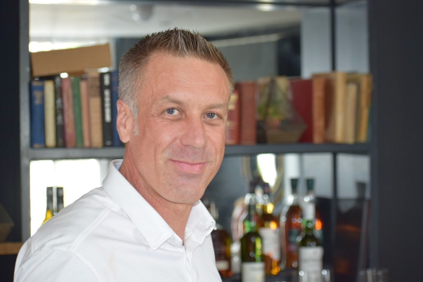 A man with short brown hair and wearing a white shirt standing in front of a bookshelf with bottles of alcohol in the background.